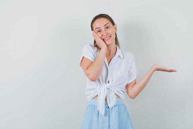 Young lady leaning cheek on palm, spreading palm in blouse and skirt and looking cute