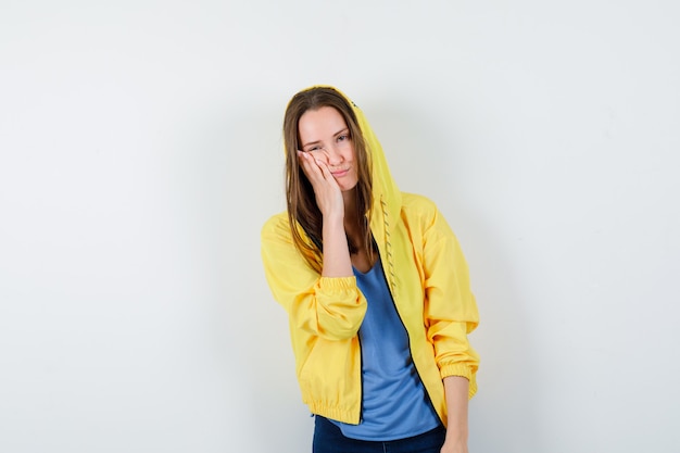 Young lady leaning cheek on palm in t-shirt, jacket and looking pensive, front view.