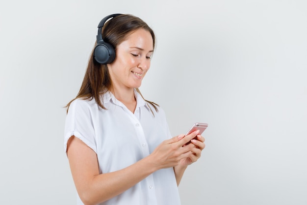 Young lady listening music while looking at her phone in white blouse and looking cheery.