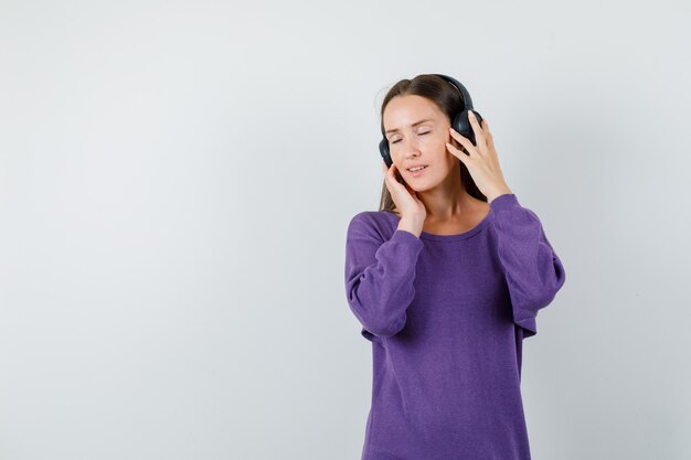 紫のシャツを着たヘッドフォンで音楽を聴いて、楽しい正面図を探している若い女性。