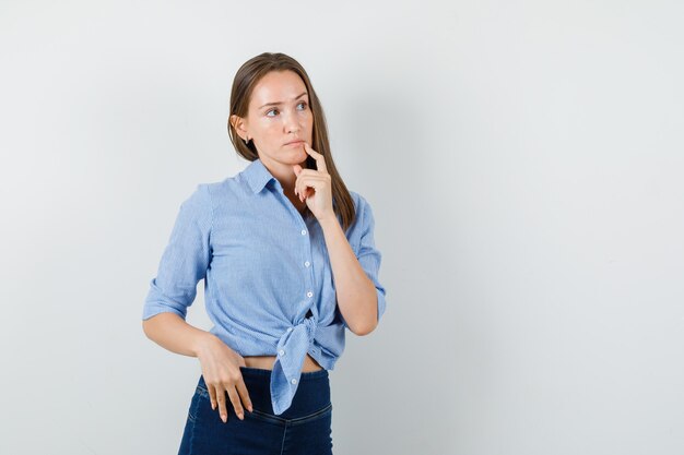 Девушка смотрит в сторону с пальцем возле рта в синей рубашке, штанах и задумчиво.