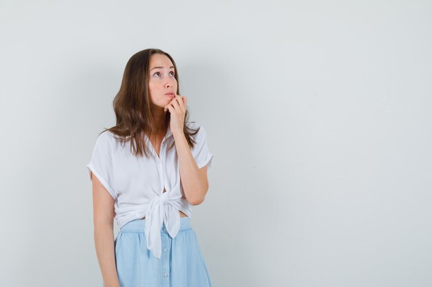 Young lady looking away with hand on chin in blouse, skirt and looking pensive