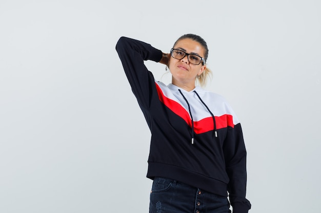 Young lady looking away with hand on neck in colorful sweatshirt,glasses front view.