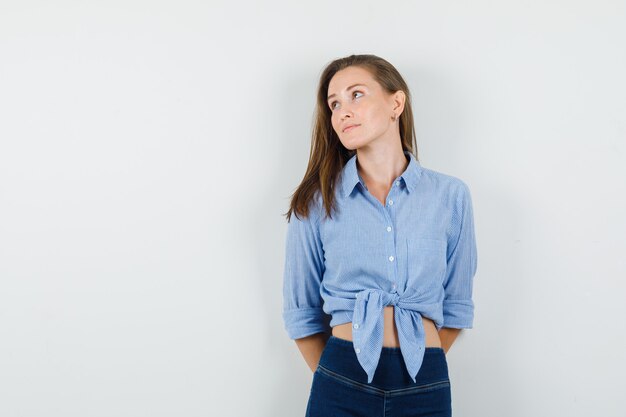 Young lady looking away with hands on her back in blue shirt, pants and looking cheery.