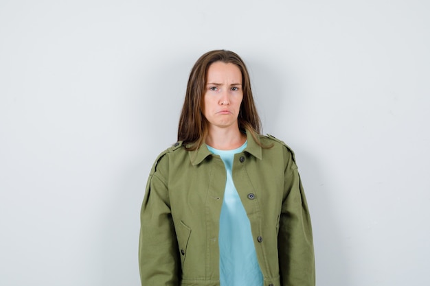 Молодая дама смотрит спереди, изогнув нижнюю губу в зеленой куртке и выглядит мрачно, вид спереди.