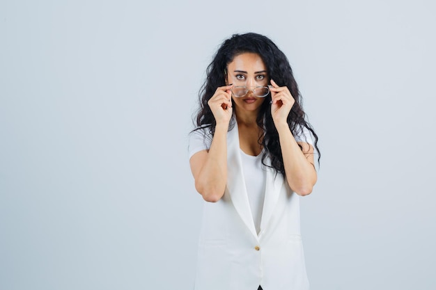 白いTシャツ、ジャケットの眼鏡を見て、疑わしい顔をしている若い女性。正面図。