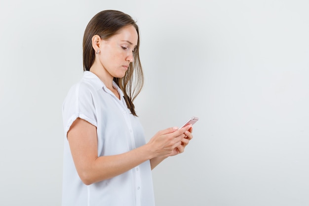 Молодая дама смотрит на свой телефон в белой блузке и выглядит грустно.
