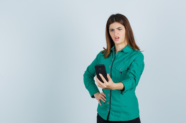 Young lady looking at mobile phone in green shirt and looking puzzled , front view.