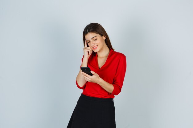 Молодая леди смотрит на мобильный телефон в красной блузке, юбке и выглядит весело