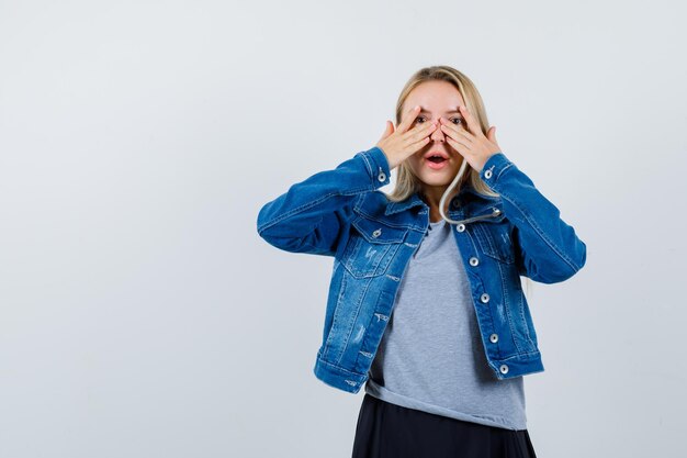 Young lady looking through fingers in t-shirt, denim jacket, skirt and looking funny