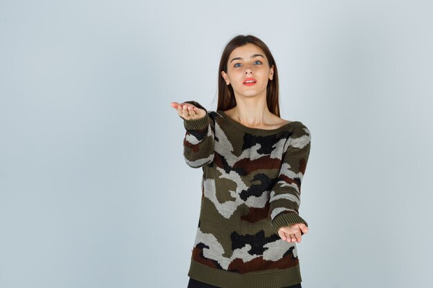 Young lady making giving gesture in sweater, skirt and looking pleased