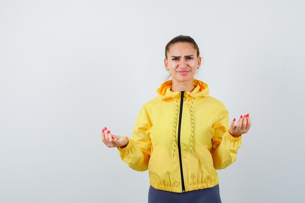 Young lady making money gesture in yellow jacket and looking displeased , front view.
