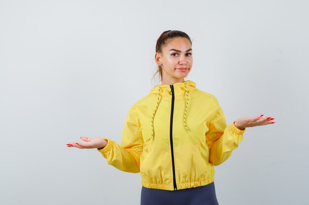 Young lady making scales gesture in yellow jacket and looking indecisive , front view.