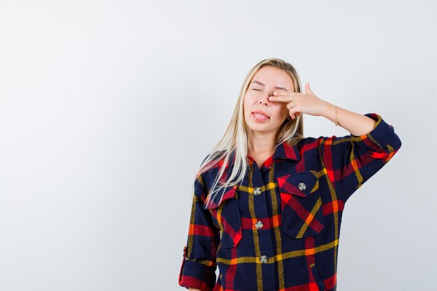 Young lady making suicide gesture while sticking out tongue in checked shirt and looking funny. front view.