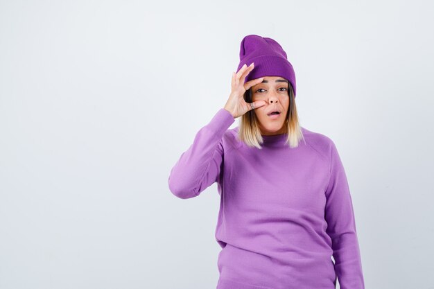 Молодая дама открывает глаз пальцем в фиолетовом свитере, шапочке и недоумевает, вид спереди.