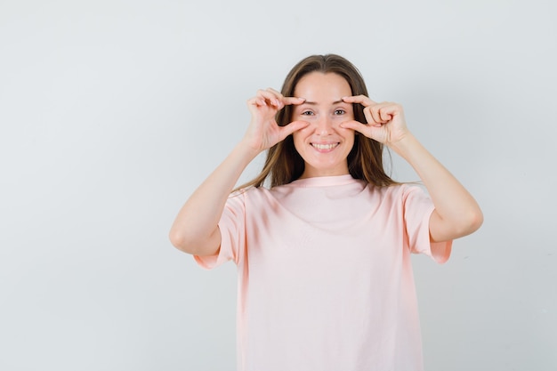 ピンクのTシャツで指で目を開けて楽観的に見える若い女性