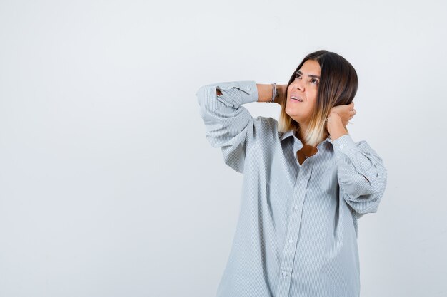 Young lady in oversize shirt holding hands behind head and looking careful , front view.