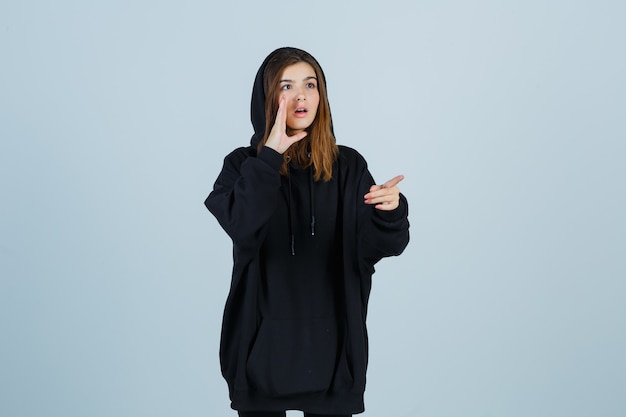 Free photo young lady in oversized hoodie, pants pointing aside while shouting something with hand and looking puzzled , front view.