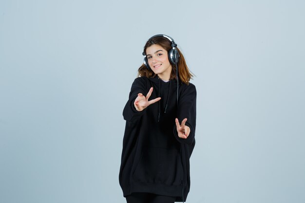 Young lady in oversized hoodie, pants showing victory sing and looking happy , front view.