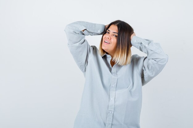 Young lady in oversized shirt holding hands on head and looking happy , front view.