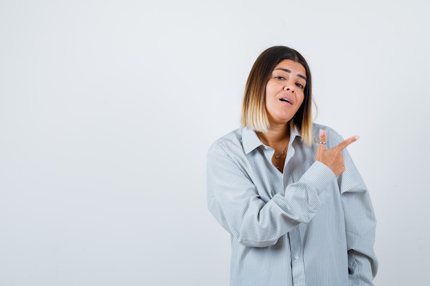 Young lady in oversized shirt pointing to the right side and looking puzzled , front view.