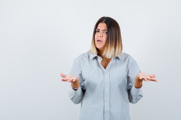 Young lady in oversized shirt spreads palms in clueless gesture and looking serious , front view.