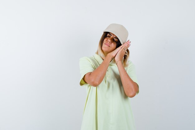 Young lady pillowing face on her hands in t-shirt, cap and looking sleepy. front view.