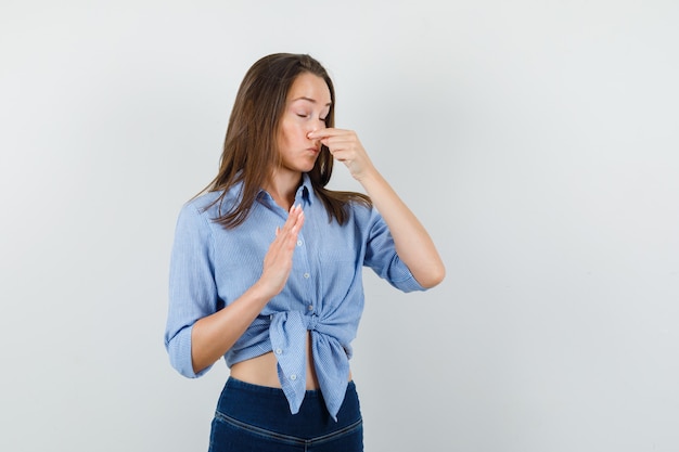 Young lady pinching nose because of bad smell in blue shirt, pants and looking disgusted