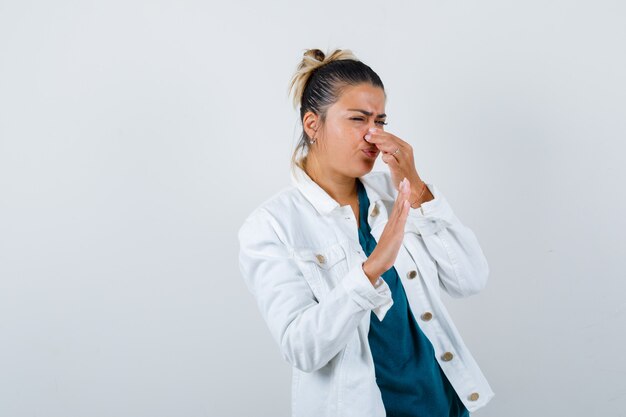 Young lady pinching nose due to bad smell in shirt, white jacket and looking disgusted , front view.