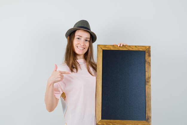 Young lady in pink t-shirt hat pointing at blackboard and looking merry  
