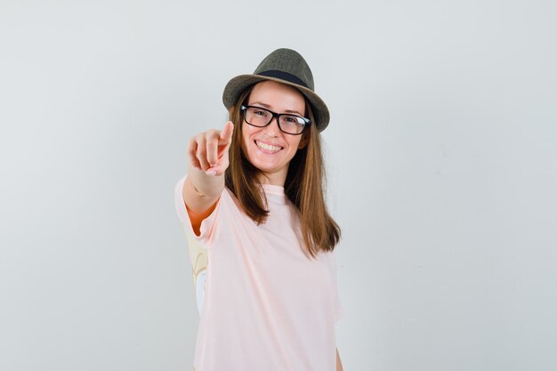 カメラを指して陽気に見えるピンクのTシャツの帽子の若い女性