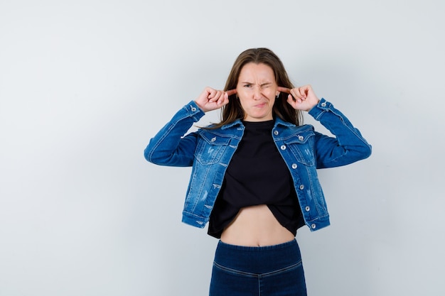 Young lady plugging ears with fingers in blouse, jacket, jeans and looking hesitant , front view.