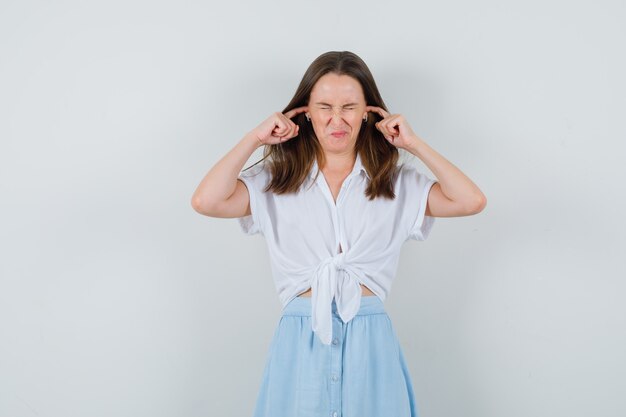 Young lady plugging ears with fingers in blouse, skirt and looking irritated