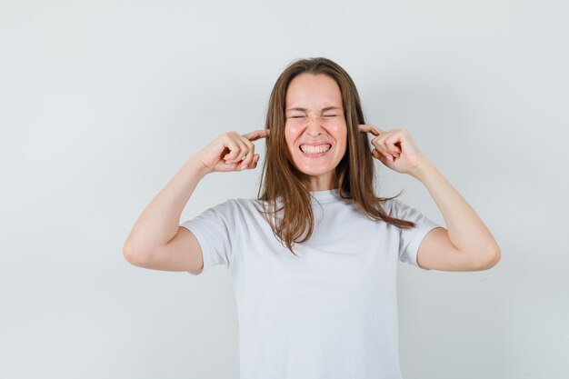 Young lady plugging ears with fingers in white t-shirt and looking annoyed 