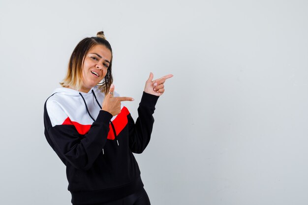 Young lady pointing aside in hoodie sweater and looking joyful.