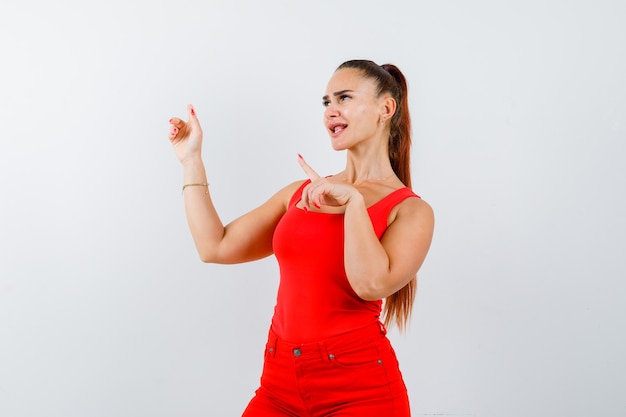 Young lady pointing aside in red singlet, red trousers and looking merry , front view.