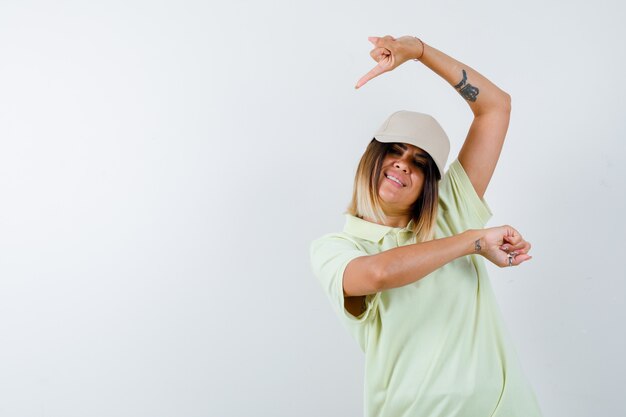 Young lady pointing aside in t-shirt, cap and looking joyful. front view.