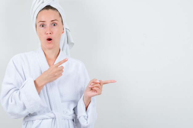 Young lady pointing aside in white bathrobe, towel and looking amazed. front view.