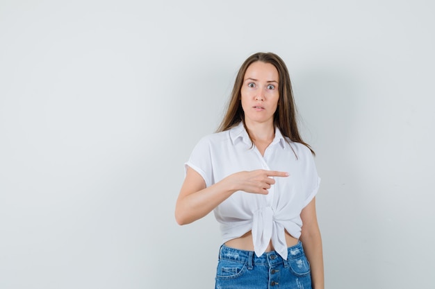 Young lady pointing aside in white blouse and looking displeased. front view.