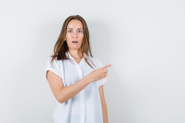 Young lady pointing aside in white blouse and looking dumbfounded