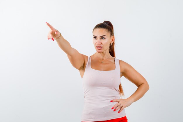 Young lady pointing away in beige tank top and looking focused , front view.