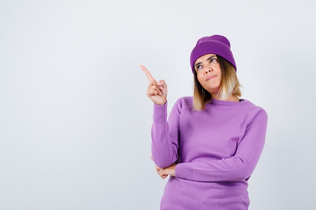 Young lady pointing away in purple sweater, beanie and looking pleased. front view.