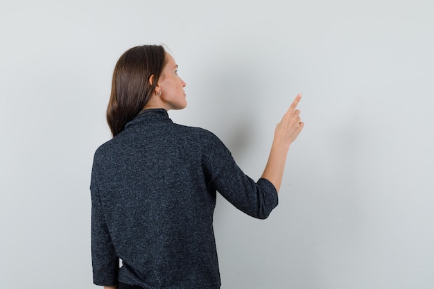 Foto gratuita giovane donna che indica lontano in camicia e guardando pensieroso, vista posteriore