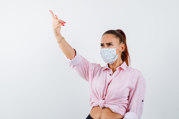 Young lady pointing away in shirt, mask and looking focused , front view.