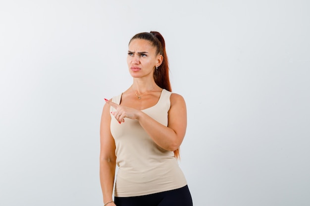 Young lady pointing away in tank top and looking pensive. front view.