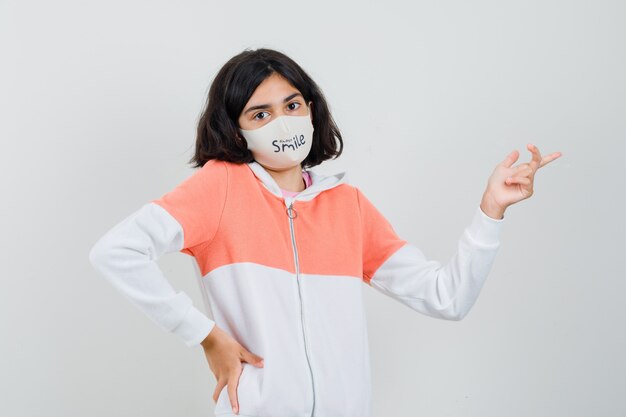 Young lady pointing away while holding hand on her waist in hoodie, face mask