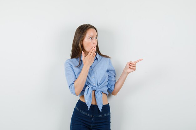 Young lady pointing away while looking aside in blue shirt, pants and looking surprised.