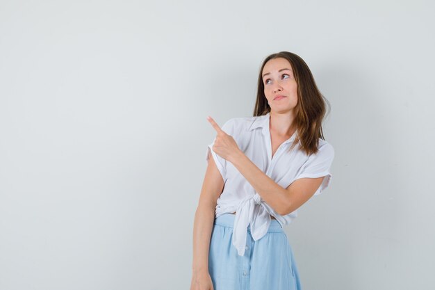 Young lady pointing away in white blouse,blue skirt and looking attentive