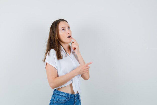 Young lady pointing away in white blouse and looking pensive. front view. space for text