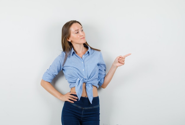 Young lady pointing away with hand on waist in blue shirt, pants and looking confident